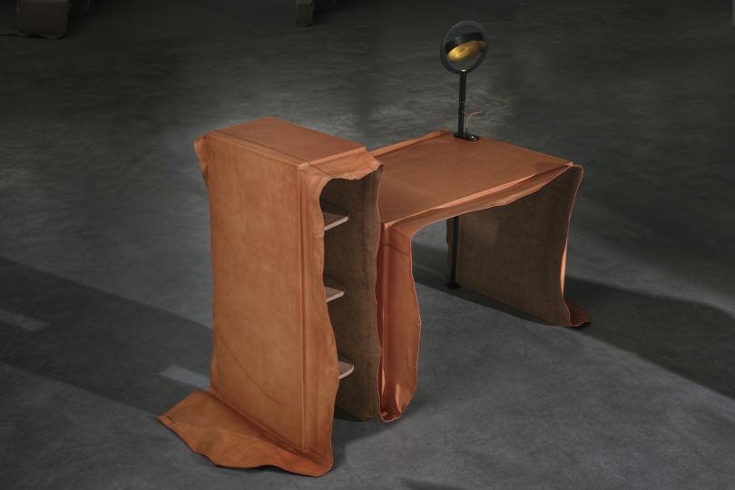 Une expo Design. Un sol en béton ciré gris foncé. Un meuble qui semble à l'envers. A gauche, une sorte d'étagère. Collée par sa droite par un plateau + une lampe. Les deux meubles semblent être recouverts d'un papier ou de cuir beige et qui déborde  de pa