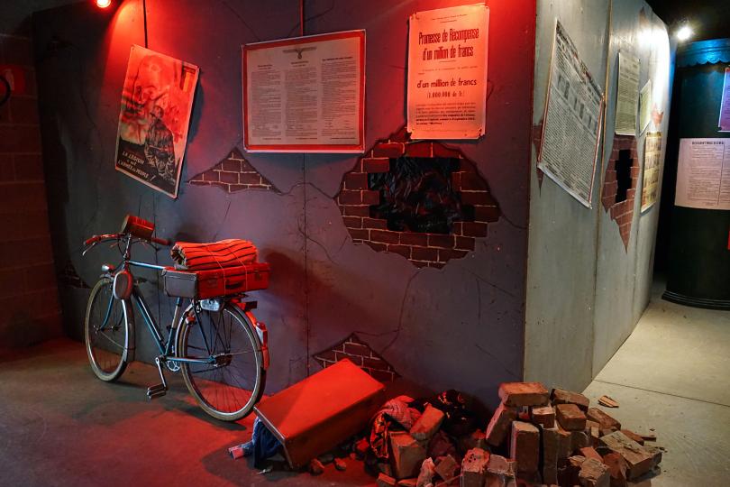 Photo d'une salle d'exposition avec un vélo et des affiches de la seconde guerre mondiale sur des murs , briques et valise à terre
