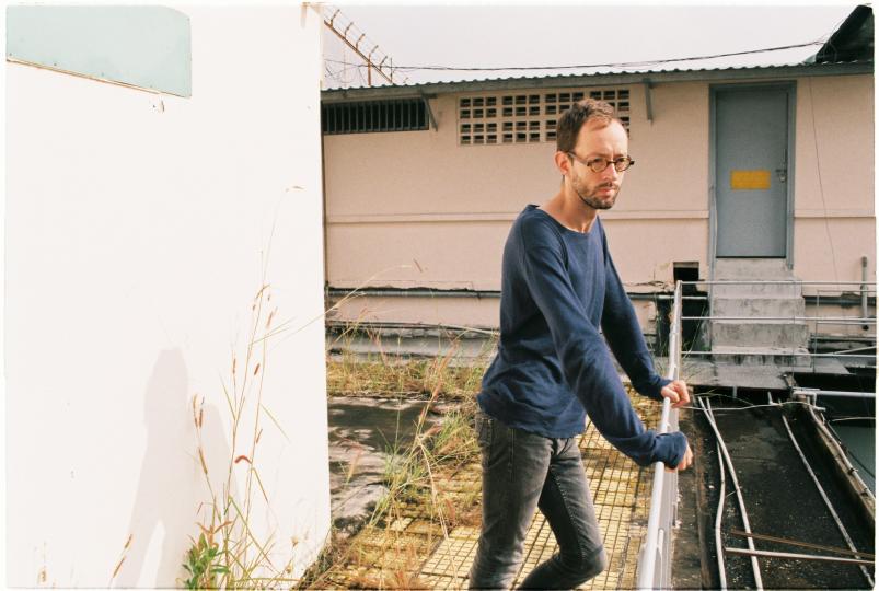 photo d'un homme à lunettes rondes les mains sur une balustrade pull bleu, des herbes mur rose