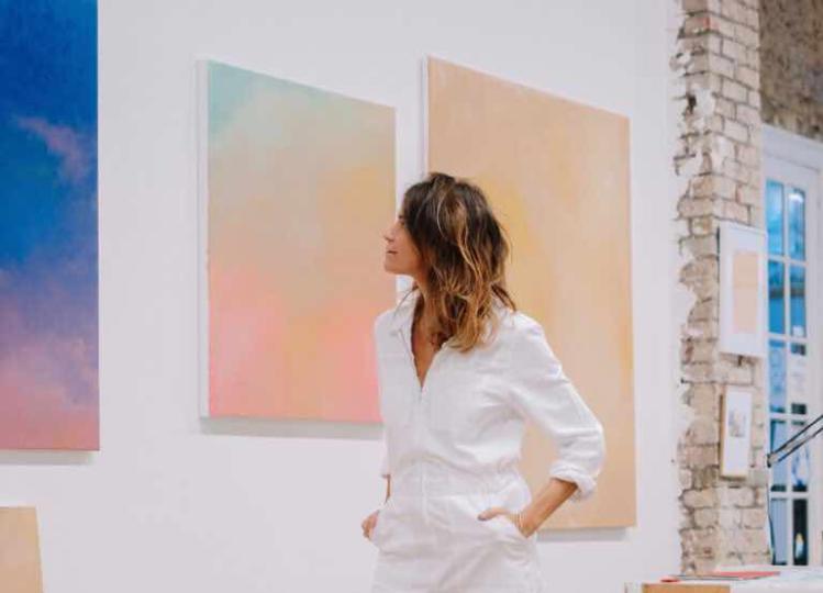 photo d'une femme en blanc devant des peintures pastel, mains dans les poches 