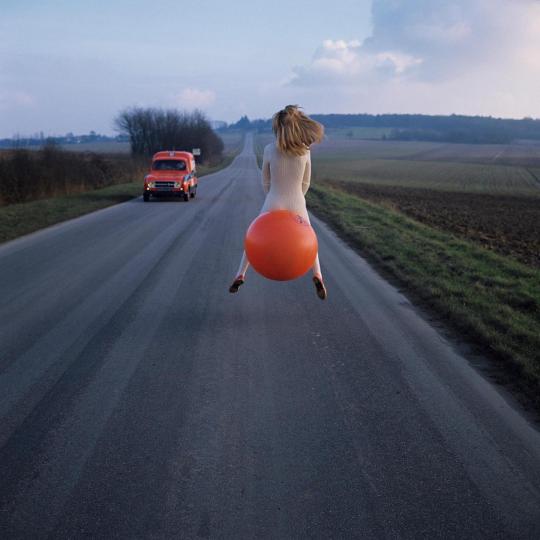 photo d'une fille de dos sur un gros ballon orange sur une route, camionnette dans l'autre sens
