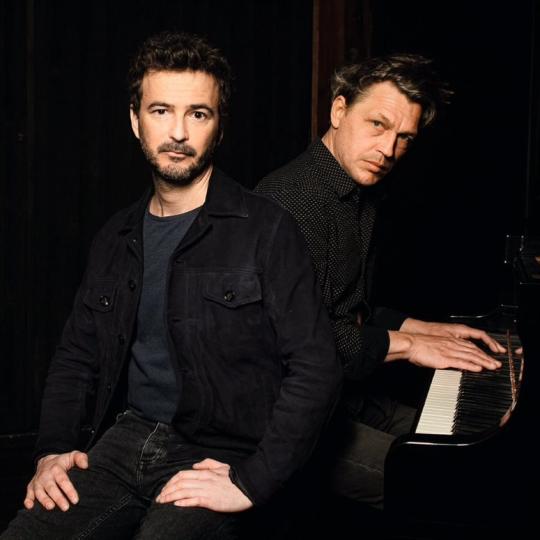 photo en noir et blanc de deux hommes assis un barbu de face et un pianiste qui regarde de son coté