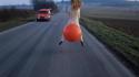 photo d'une fille de dos sur un gros ballon orange sur une route, camionnette dans l'autre sens