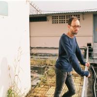 photo d'un homme à lunettes rondes les mains sur une balustrade pull bleu, des herbes mur rose